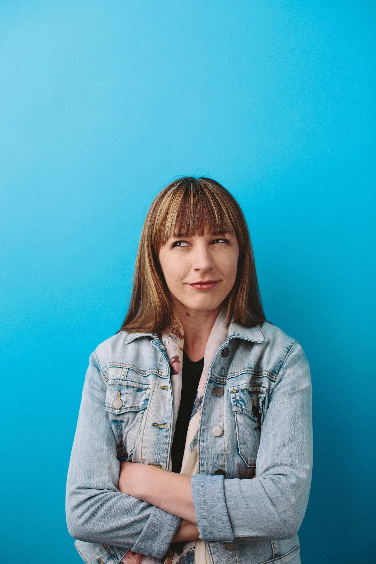 A woman wearing a denim jacket looking thoughtfully off-camera against a bright blue studio background.