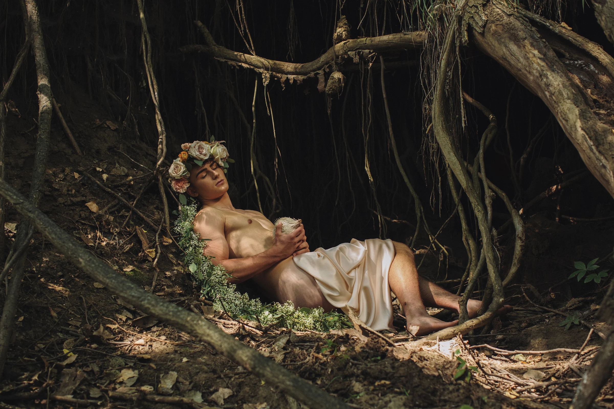 A man wearing a floral crown and draped in white fabric reclining on a large, twisting tree branch in a forest setting.